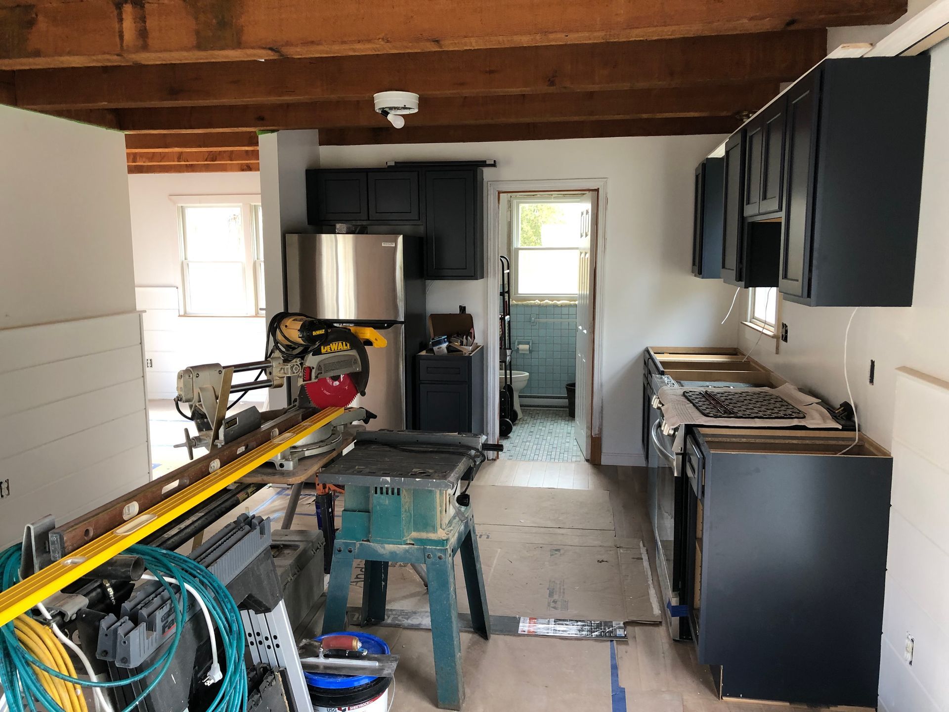 Narrow kitchen under renovation with exposed beams, cabinets, tools, and appliances along both sides.