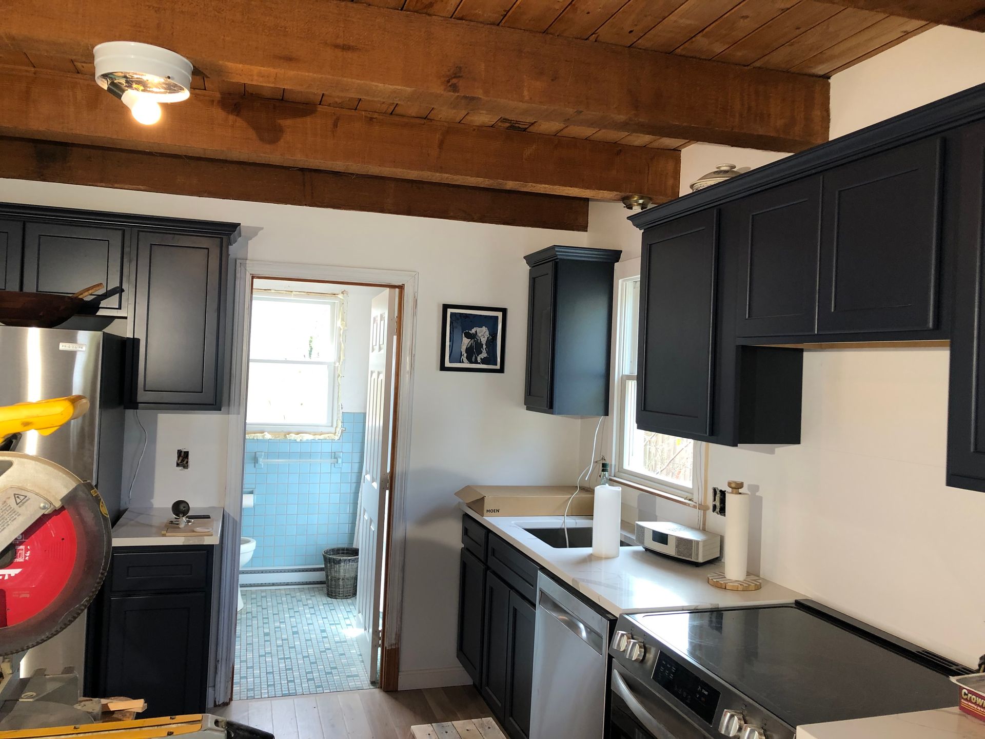 Small kitchen with black cabinets, white counters, wood ceiling, and an open doorway to a blue-tiled bathroom.
