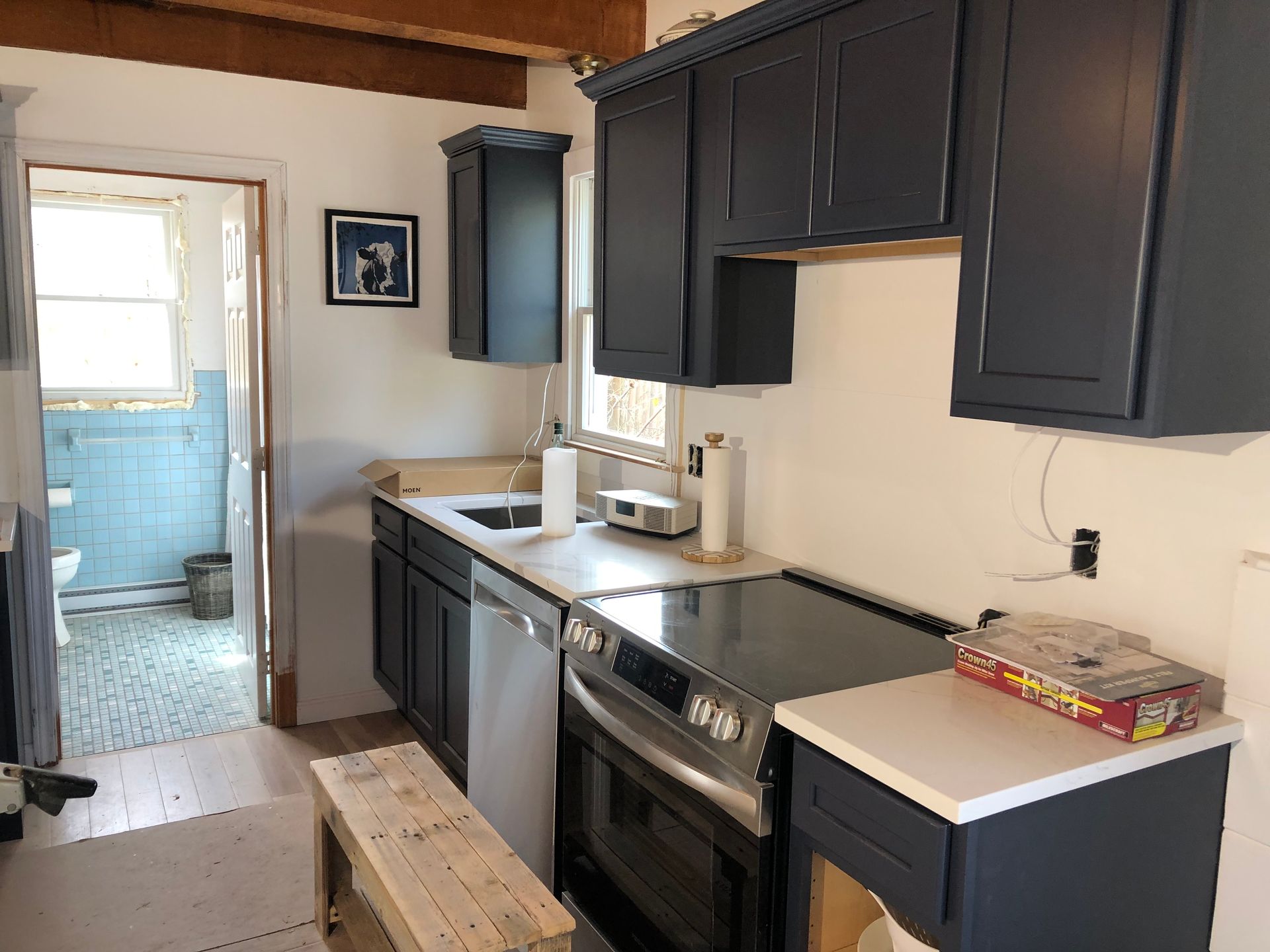 Narrow kitchen with dark cabinets, white counters, a stove, sink, and doorway to a blue-tiled room