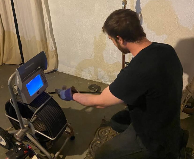 A person using a drain inspection camera in a basement. The camera displays a blue screen while they work.