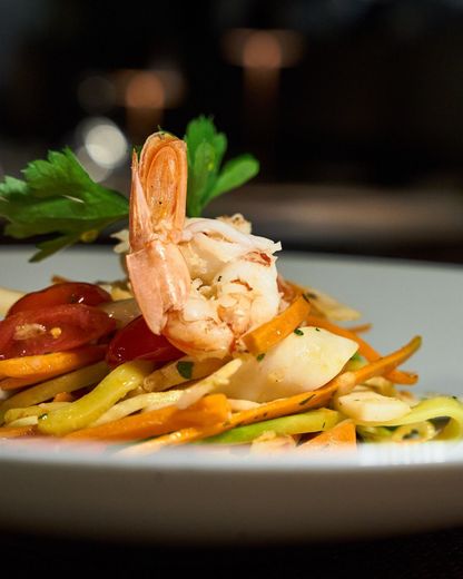 Pasta con gamberi, pomodorini, verdure gialle e prezzemolo, servita su un piatto bianco.