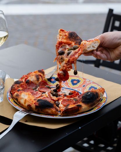 Una mano solleva una fetta di pizza al salame piccante da un piatto a un tavolo all'aperto di un ristorante.
