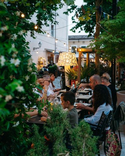 Persone sedute ai tavoli nel patio di un caffè all'aperto immerso nel verde, illuminate da lucine, al crepuscolo.