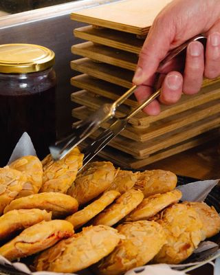 Pinze da cucina che sollevano pasticcini dorati da un vassoio accanto a teglie da forno impilate e un barattolo di marmellata.