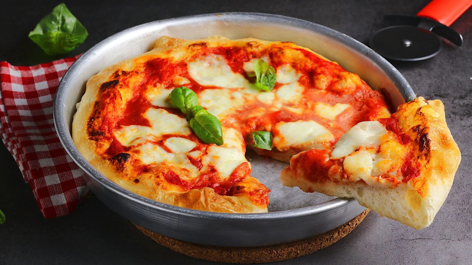 Pizza al formaggio in una teglia di metallo con foglie di basilico, una fetta viene sollevata su un tavolo scuro