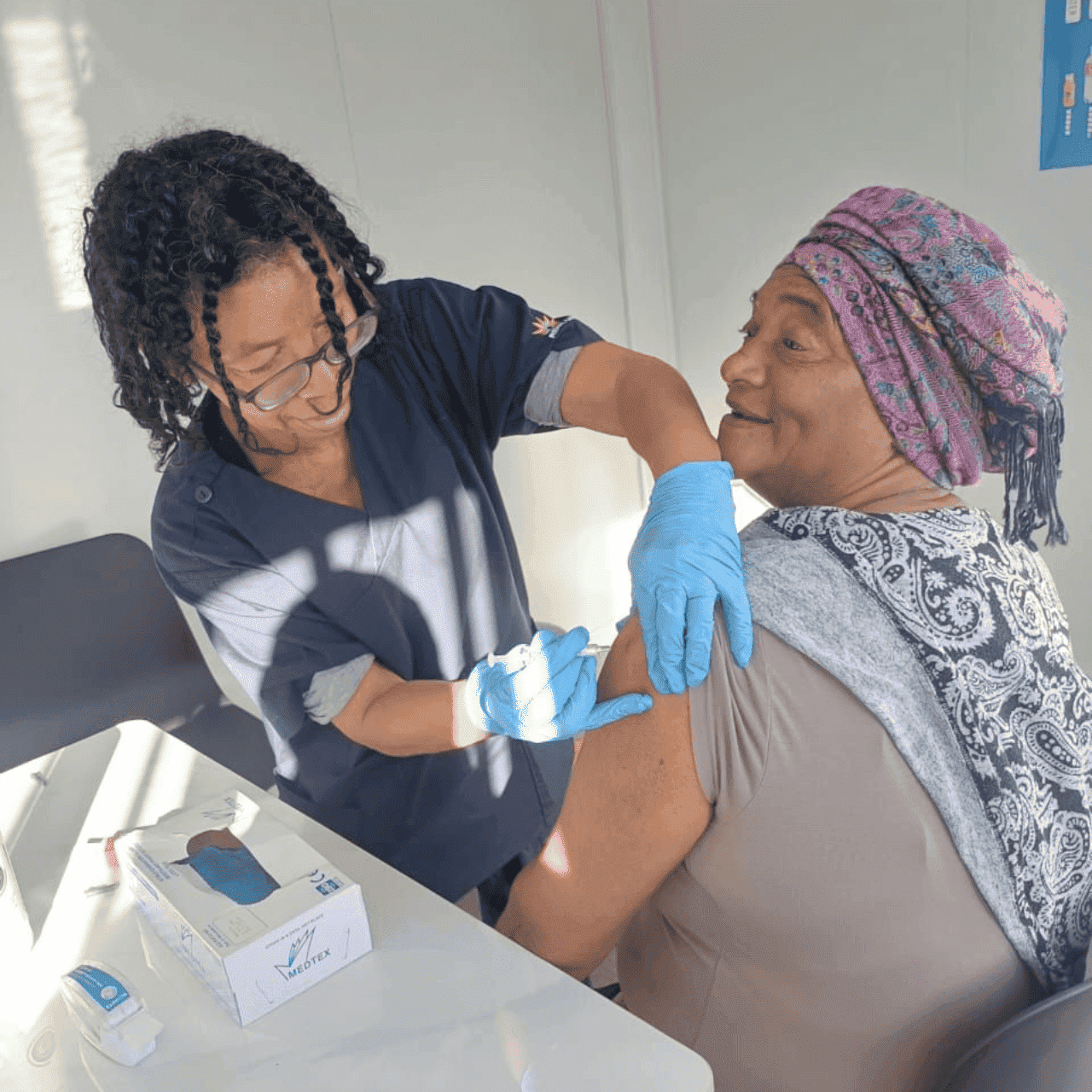 Healthcare worker giving an injection to a seated patient indoors, both wearing gloves.