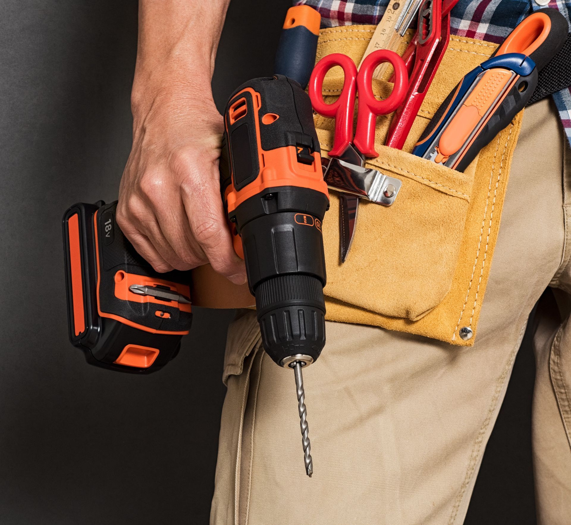 Hand holding an orange-and-black cordless drill with tool belt of screwdrivers and pliers on khaki pants