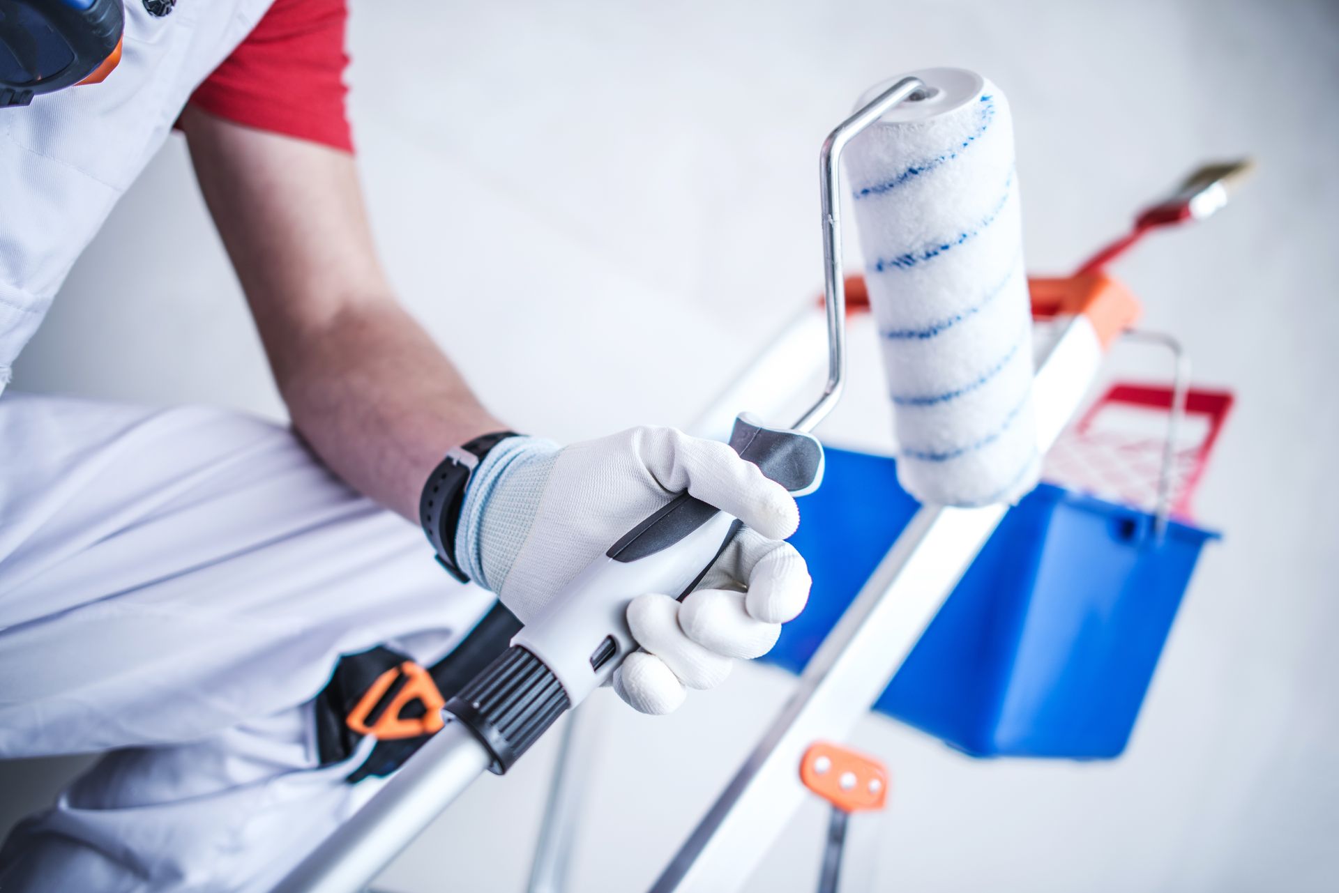 Gloved hand painting a white railing with a brush, with a blue bucket and red ladder in the background
