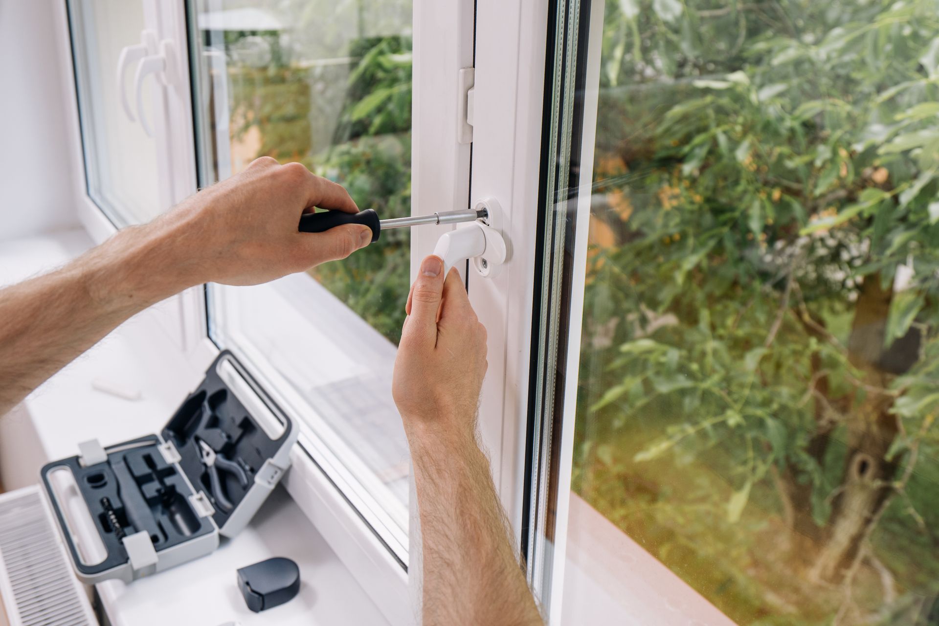 Hands using a screwdriver to adjust a white window hinge beside an open tool case.