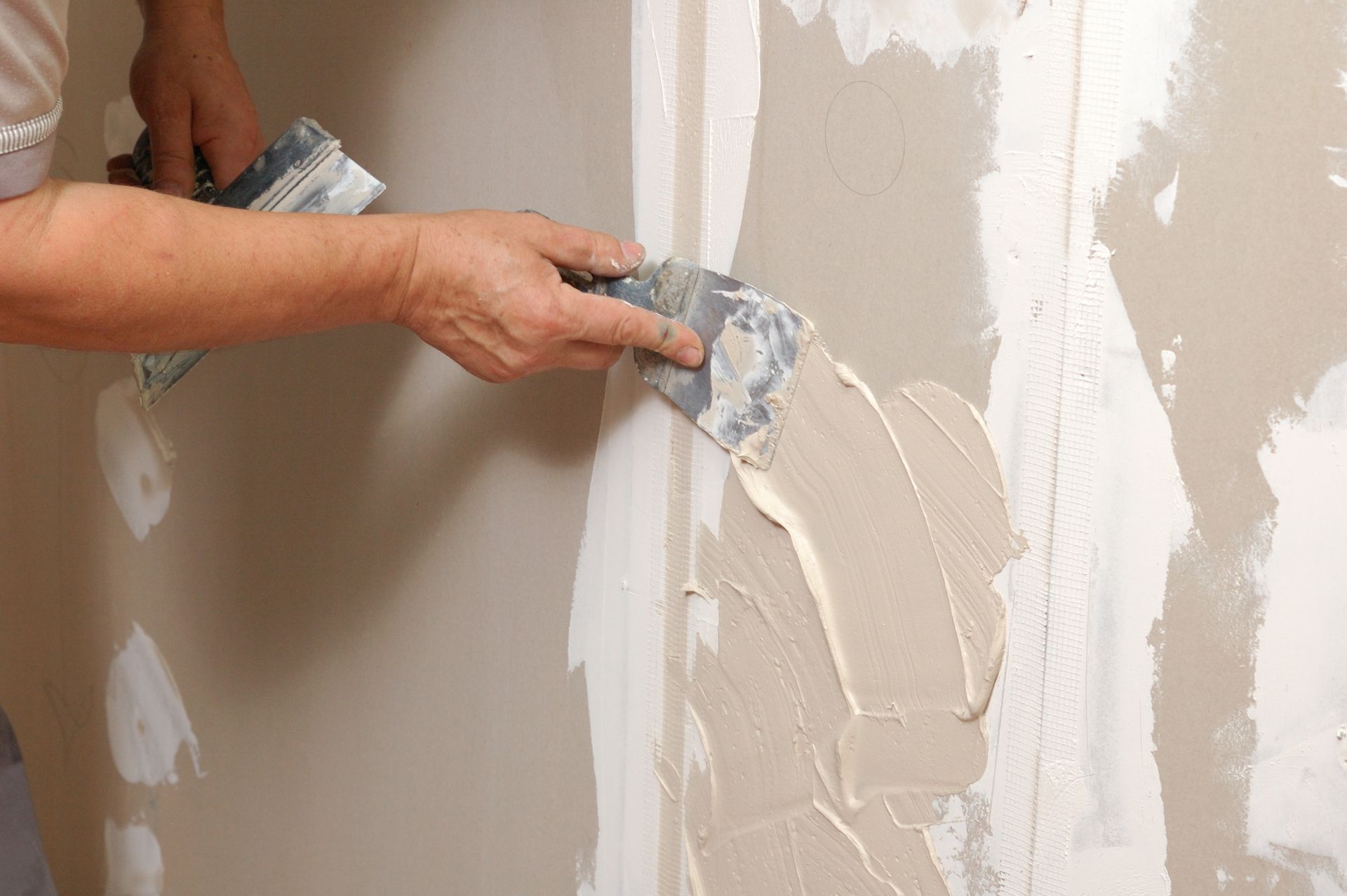 Hands applying joint compound to a beige wall with a putty knife during drywall repair