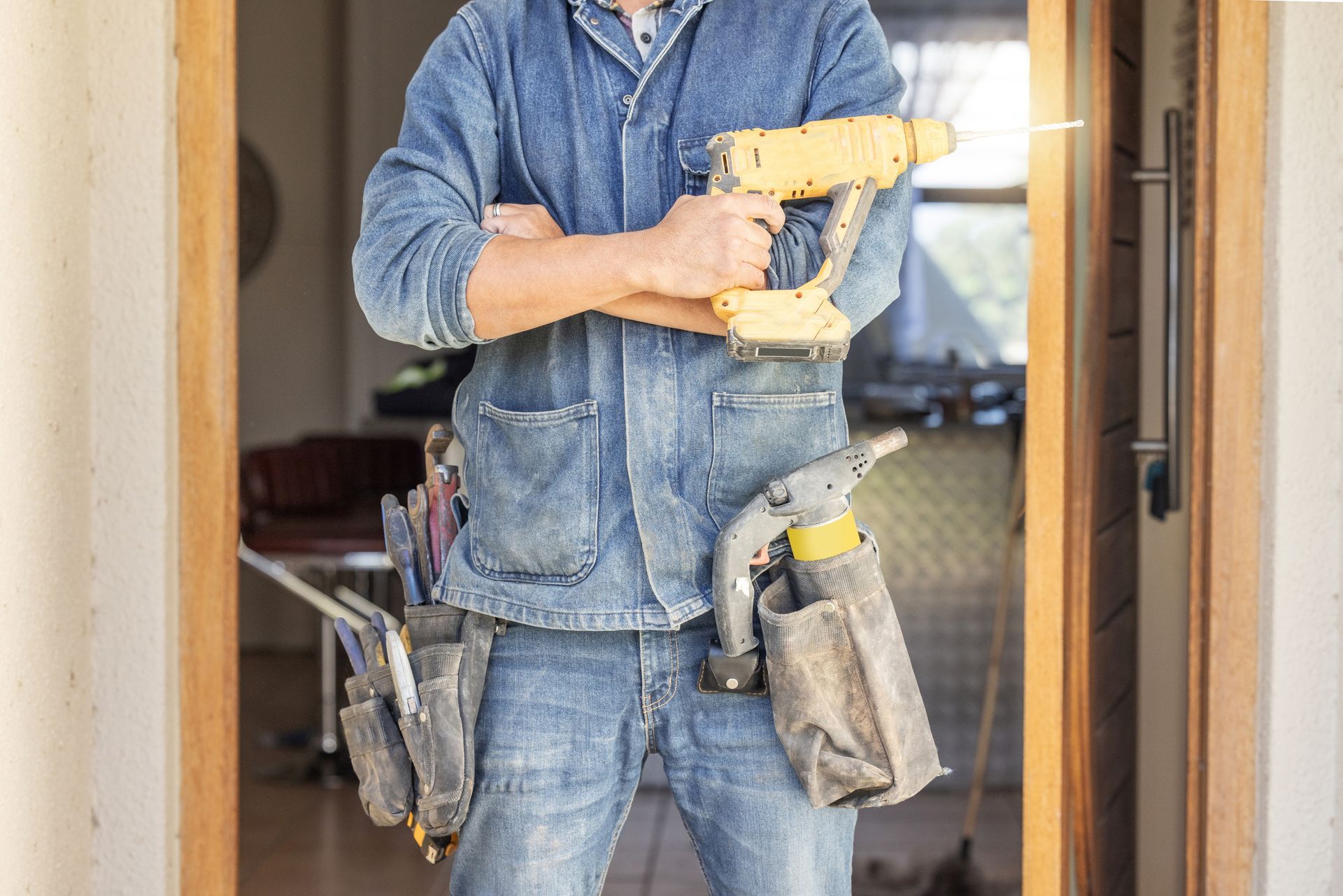Worker in blue denim holding a cordless drill with tool pouches in a doorway