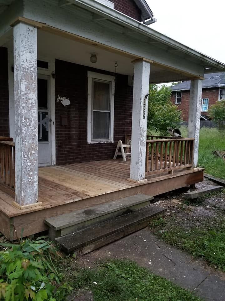 Front porch of a weathered house with peeling white columns, wooden deck, and steps leading down to a walkway