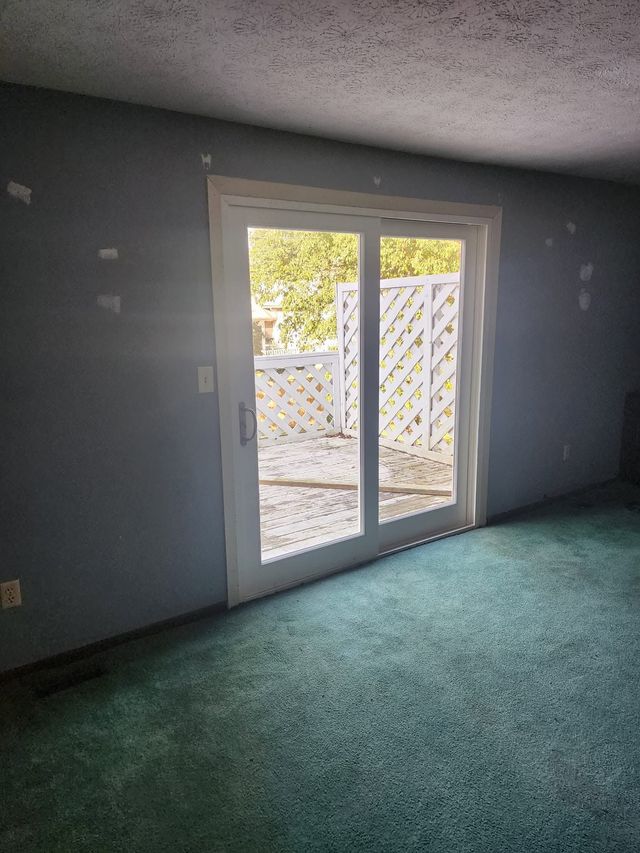 Empty room with gray walls, green carpet, and sliding glass doors opening to a fenced patio.