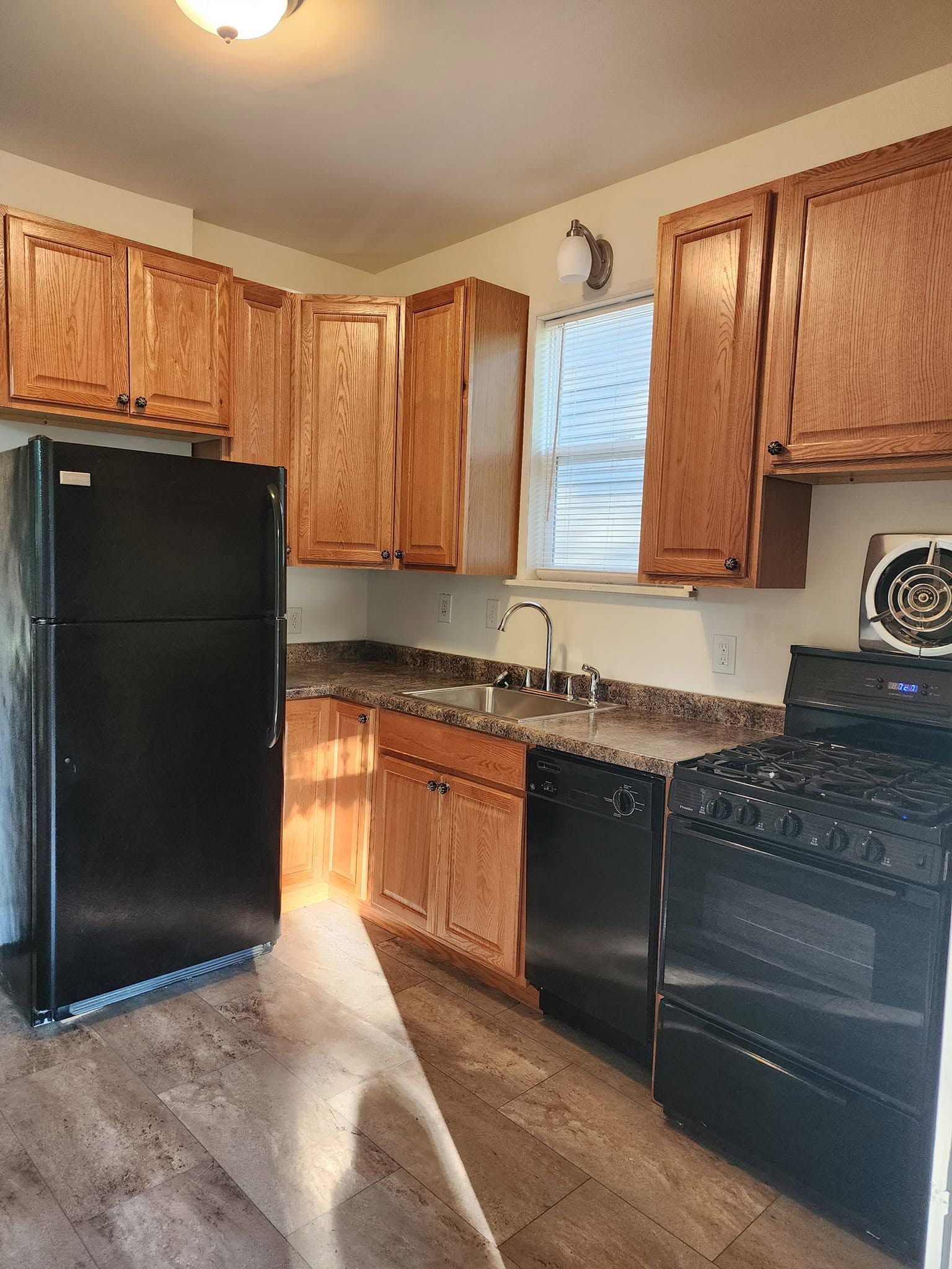 Small kitchen with wood cabinets, black appliances, tiled floor, and sunlight through the window.