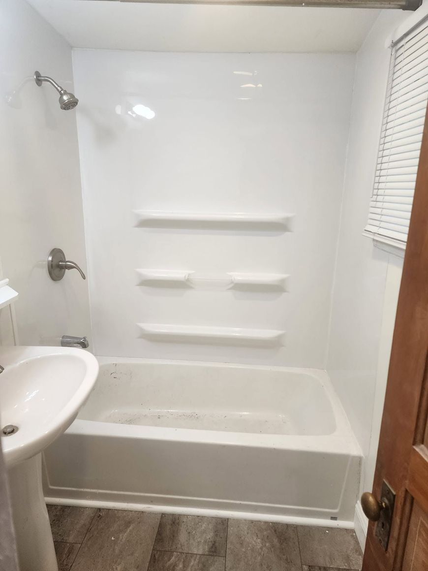 Small white bathroom with tub-shower combo, built-in shelves, sink, and window blinds.