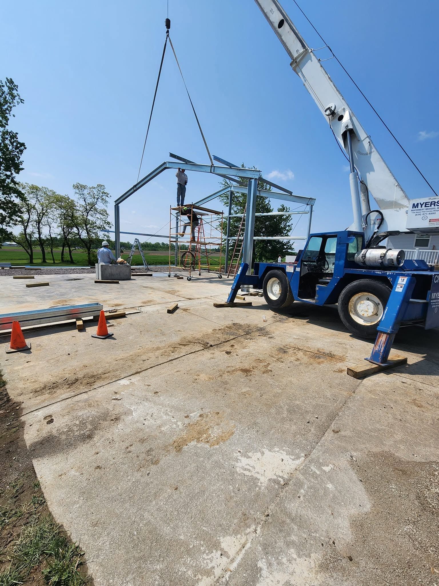 Blue crane lifting a steel frame on a concrete lot under a clear sky