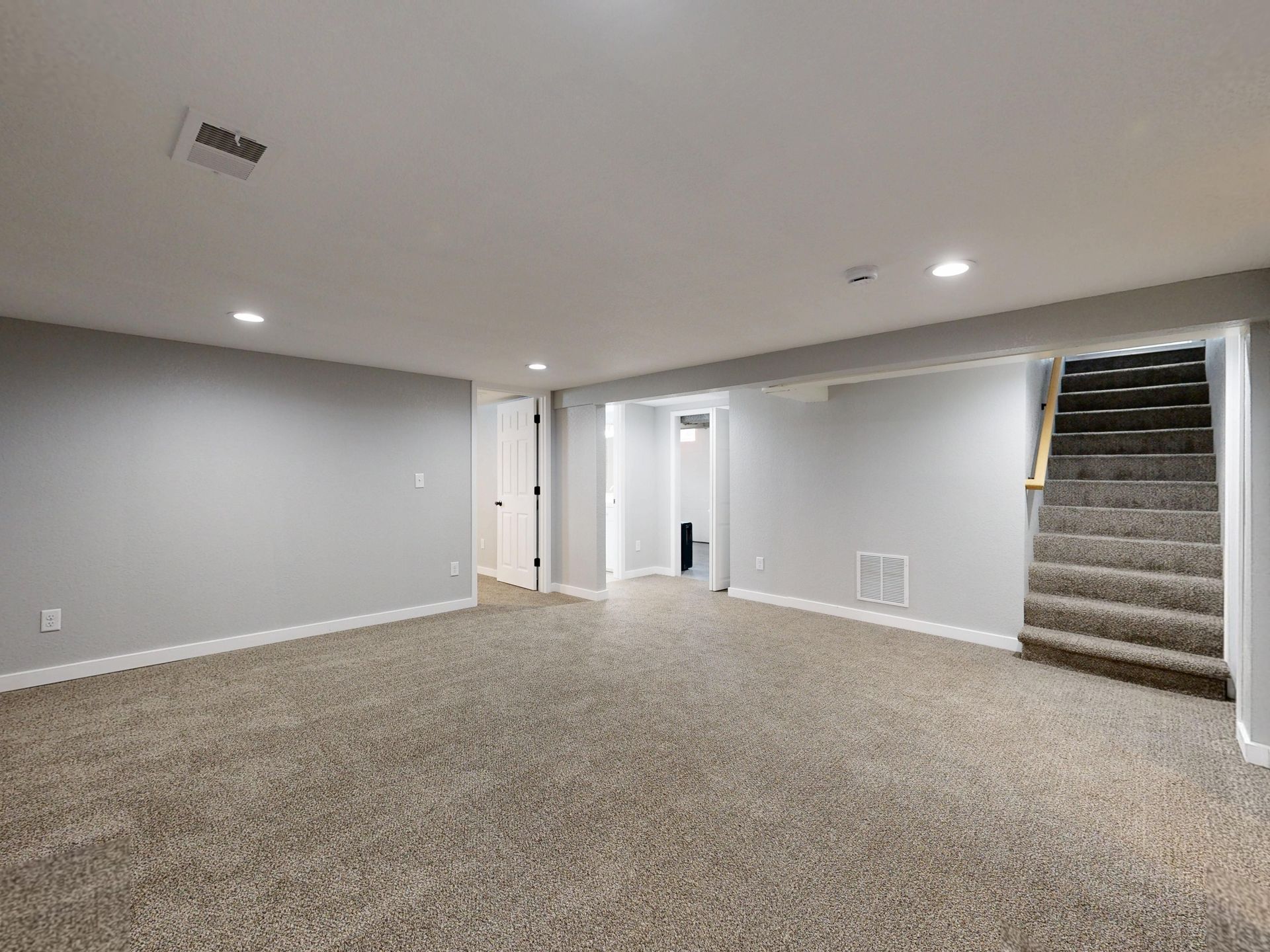 Empty carpeted basement room with gray walls, recessed lights, and stairs leading up on the right.