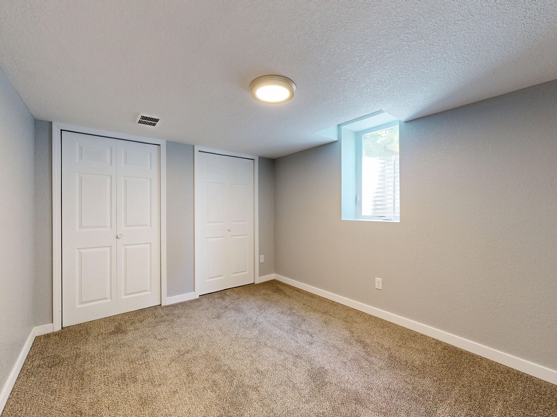 Empty beige bedroom with carpet, two white closet doors, and a small window on gray walls