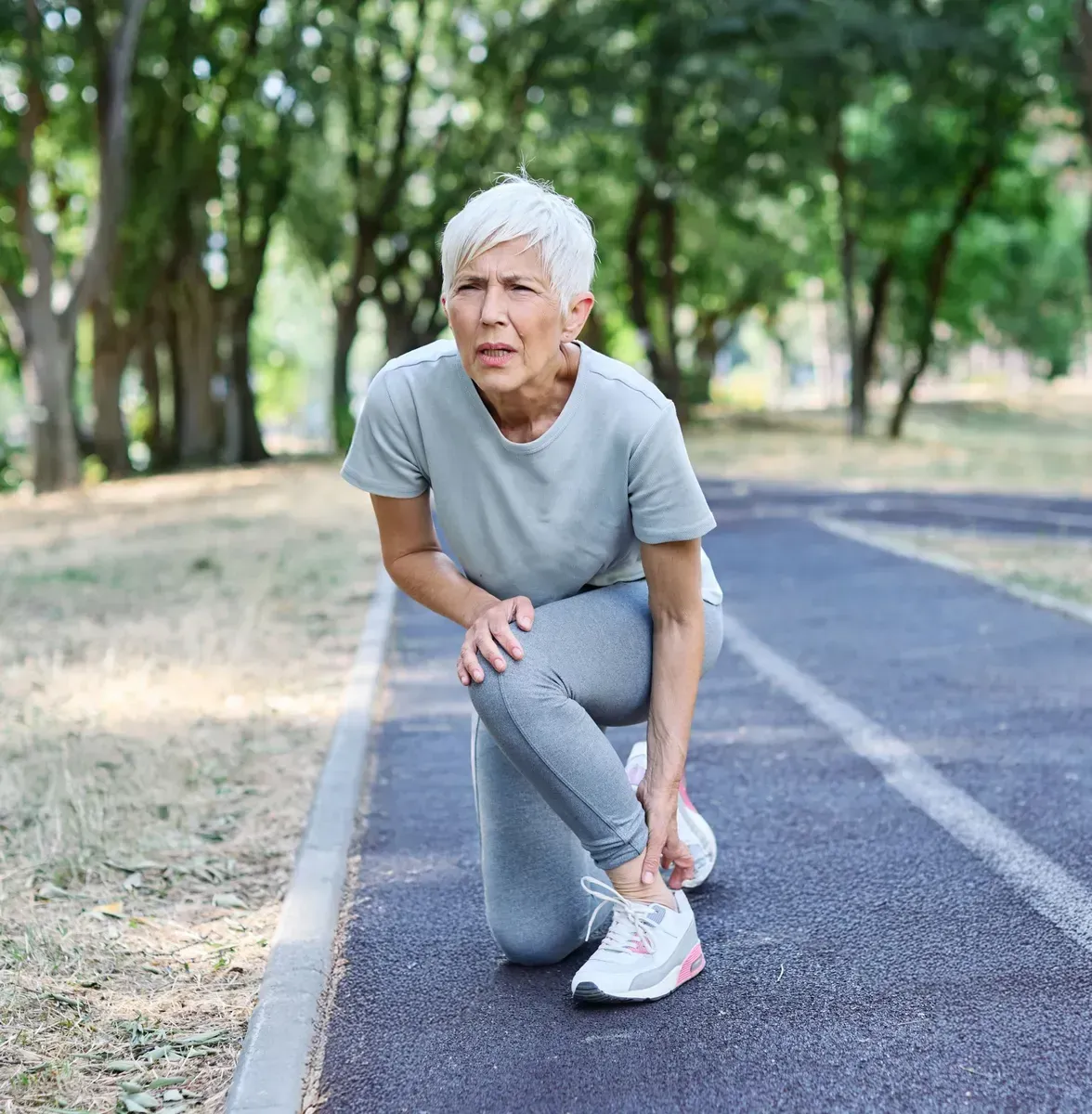 Older runner in gray clothes pauses to adjust shoe on a park running path