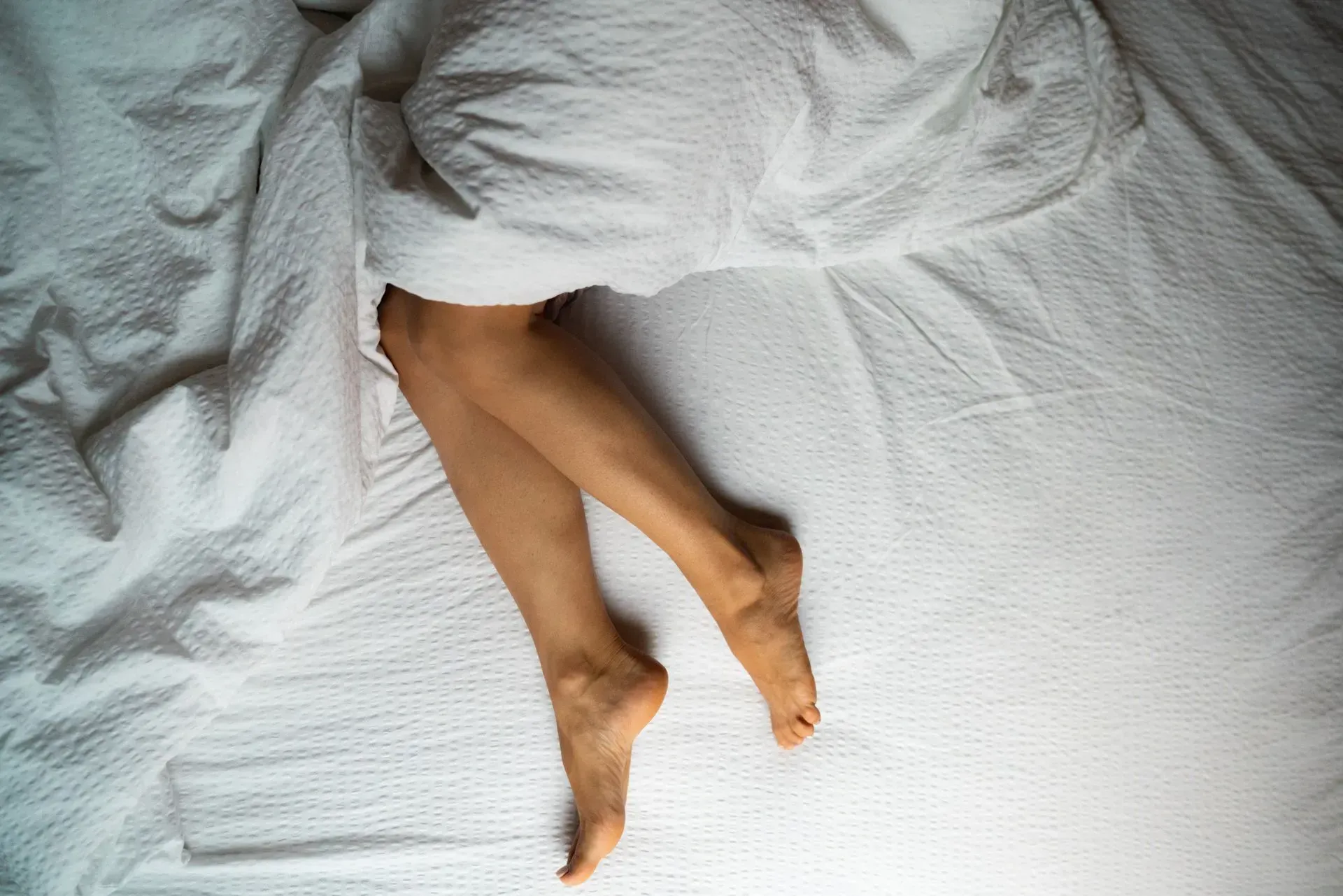 Legs crossing on a white bed under a rumpled white blanket