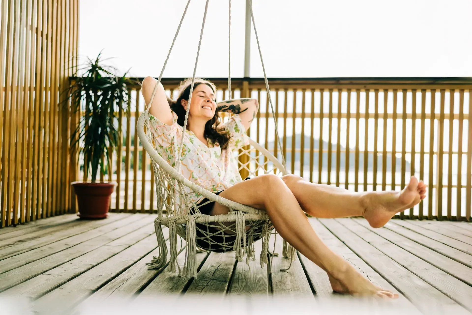 Person relaxing in a woven chair on a wooden deck, smiling with legs outstretched in warm sunlight.