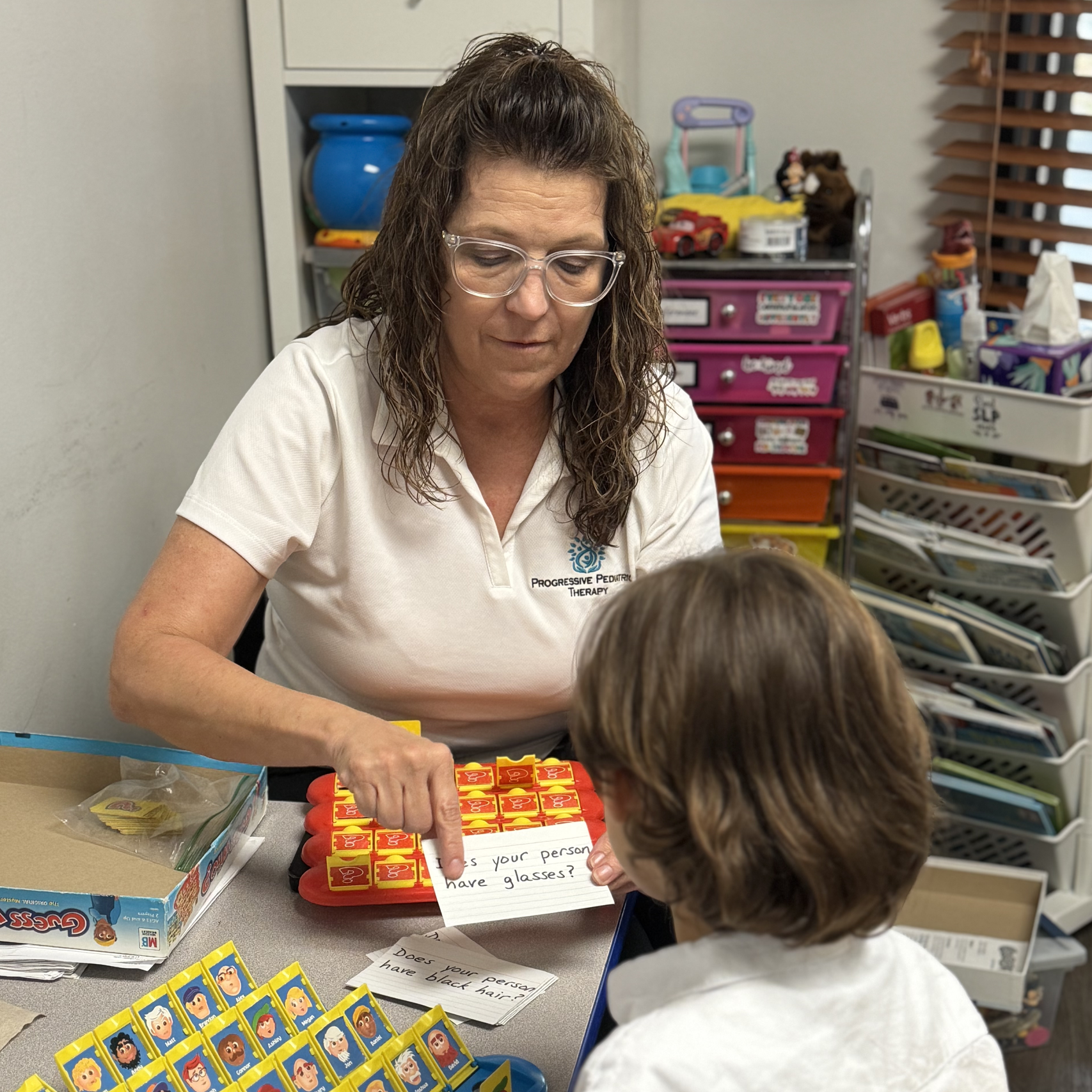 Woman points at card during Guess Who game with child, in a room with toys and shelves.