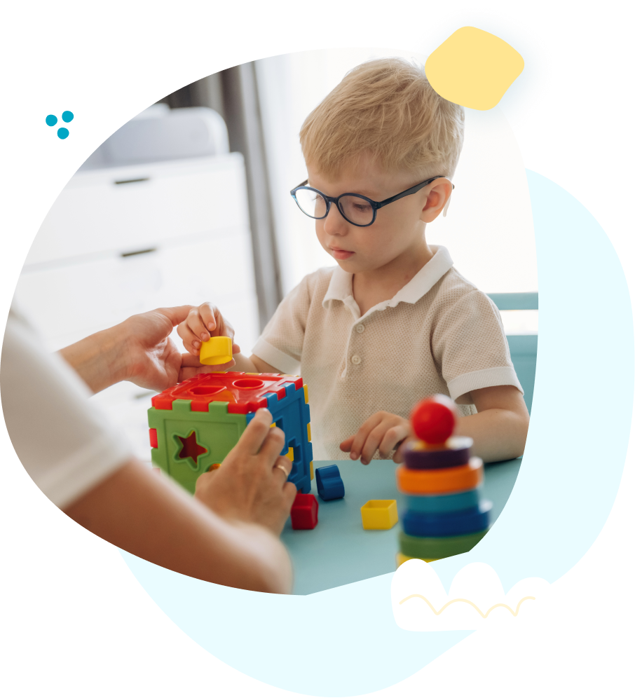Child with glasses playing with colorful toys at a table with an adult's hands nearby