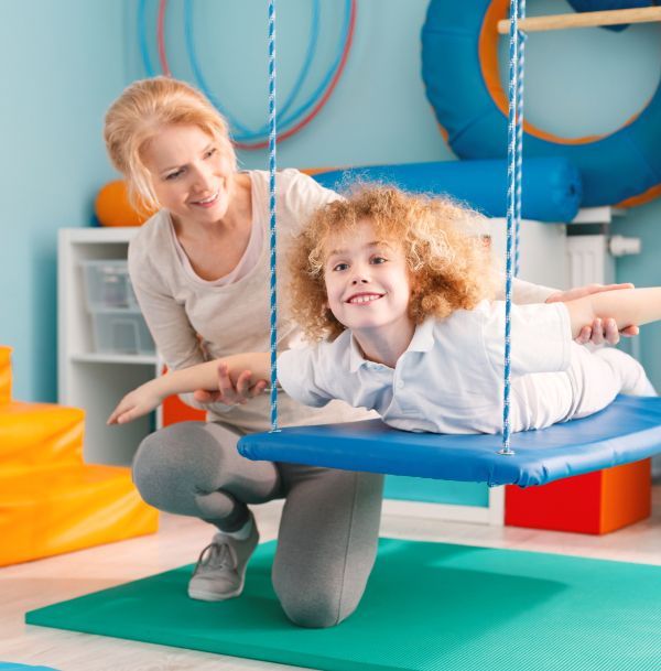 Woman assisting a child on a blue swing in a therapy room, child smiling, arms outstretched.