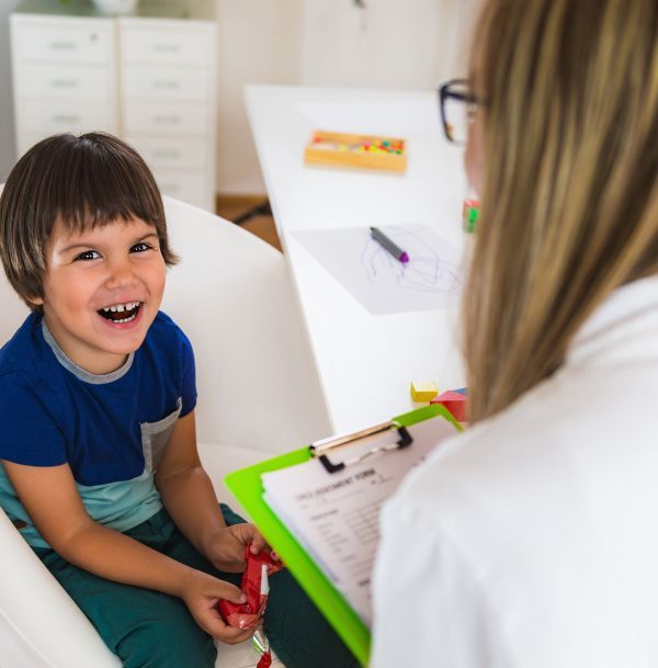Boy smiling at therapist holding clipboard in a brightly lit room.