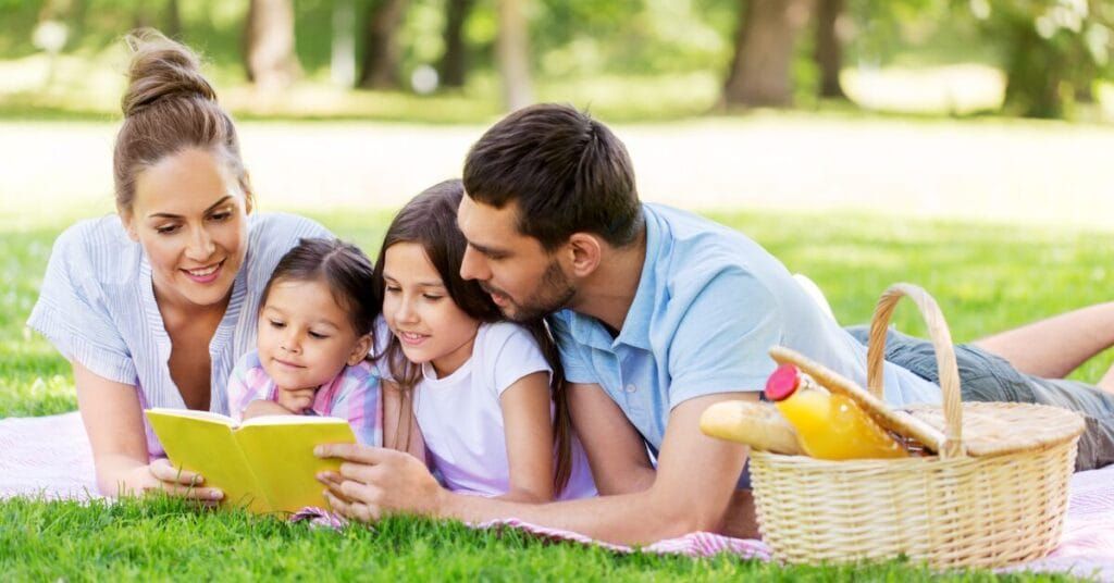 A family is laying on a blanket in the grass reading a book.