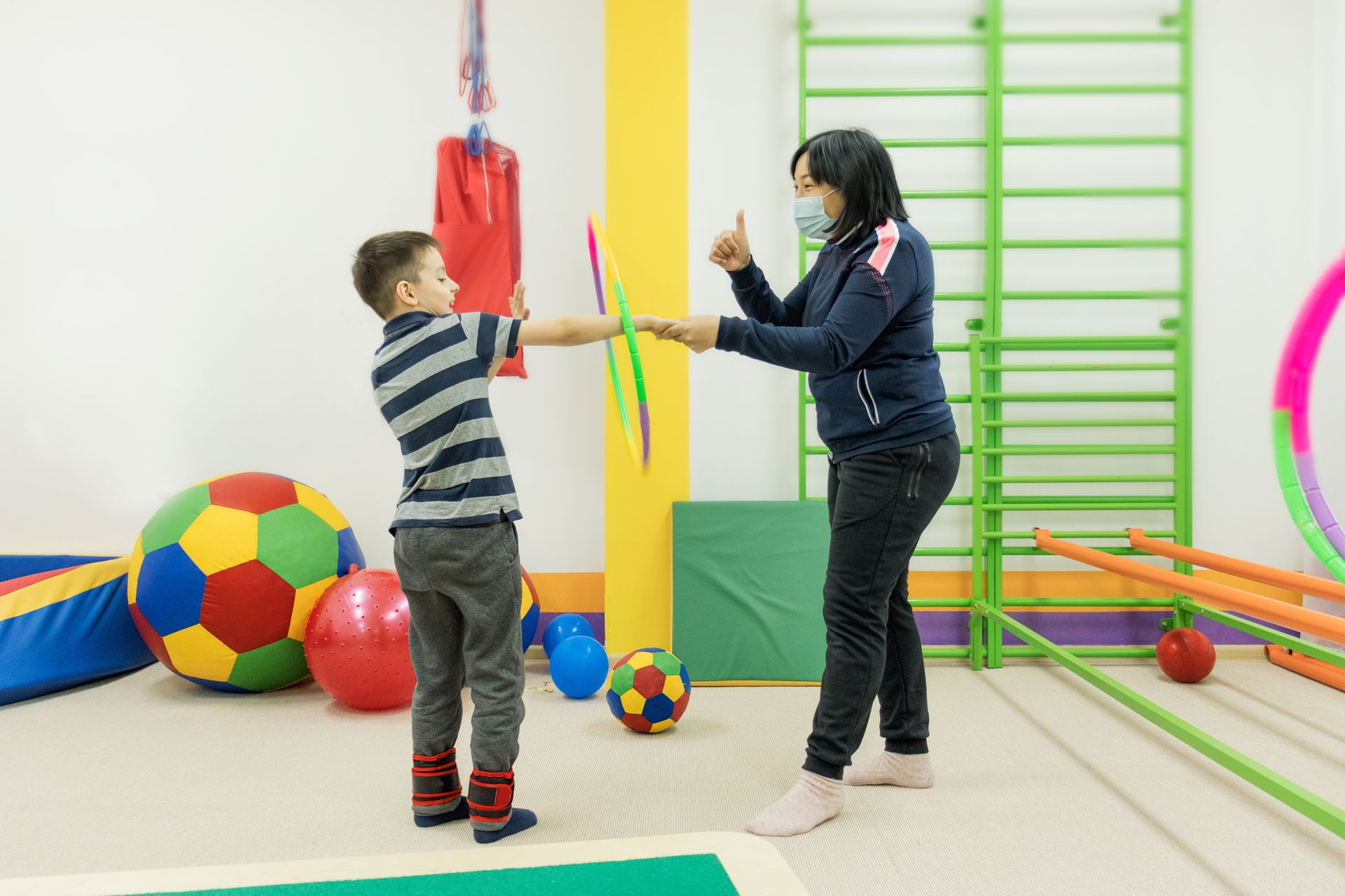 Two people reaching toward a rainbow ribbon in a colorful gym with exercise balls and climbing bars