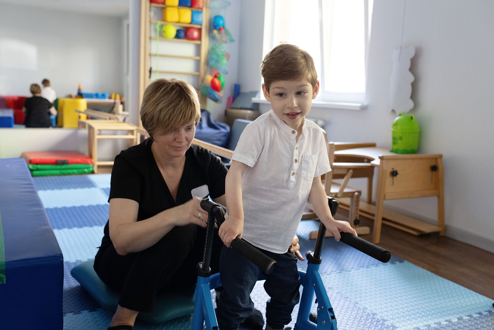 Adult assisting child in a blue mobility walker in a bright playroom