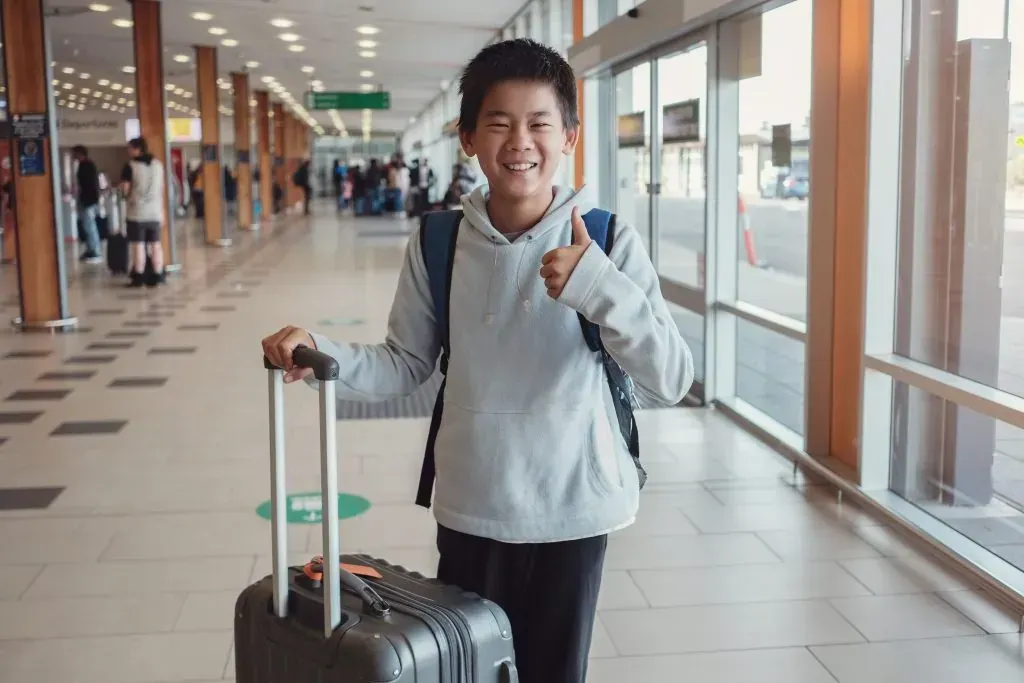 A young boy is walking with a suitcase at an airport and giving a thumbs up.