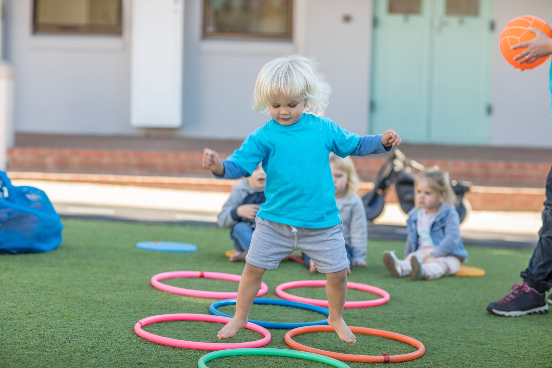 Child in a blue shirt stepping through colorful hoops on grass while other children watch nearby