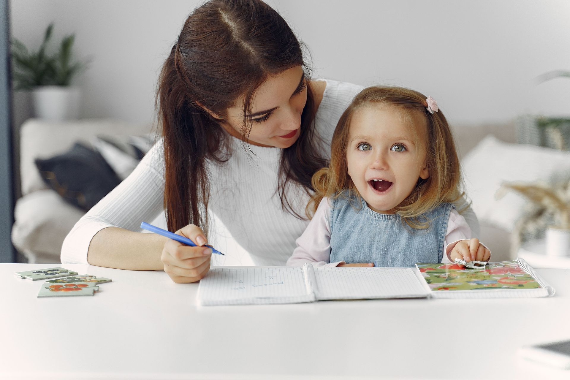 Woman helping a child draw at a white table, with the child smiling in a bright room.