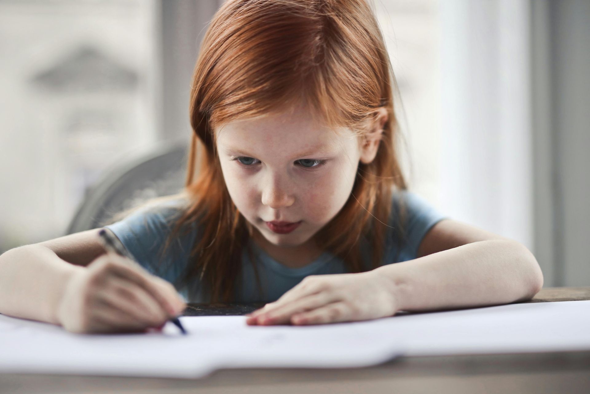 Girl with red hair writing on white paper at a table.