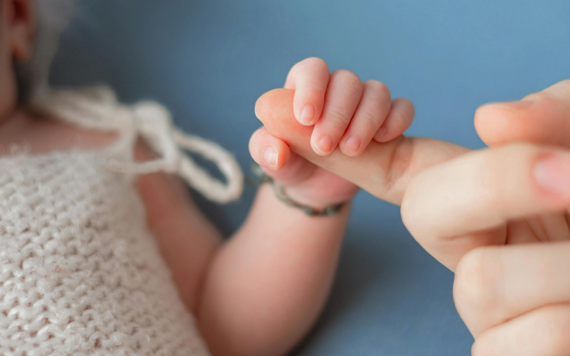 Baby's tiny hand holding an adult's finger against a light blue background.
