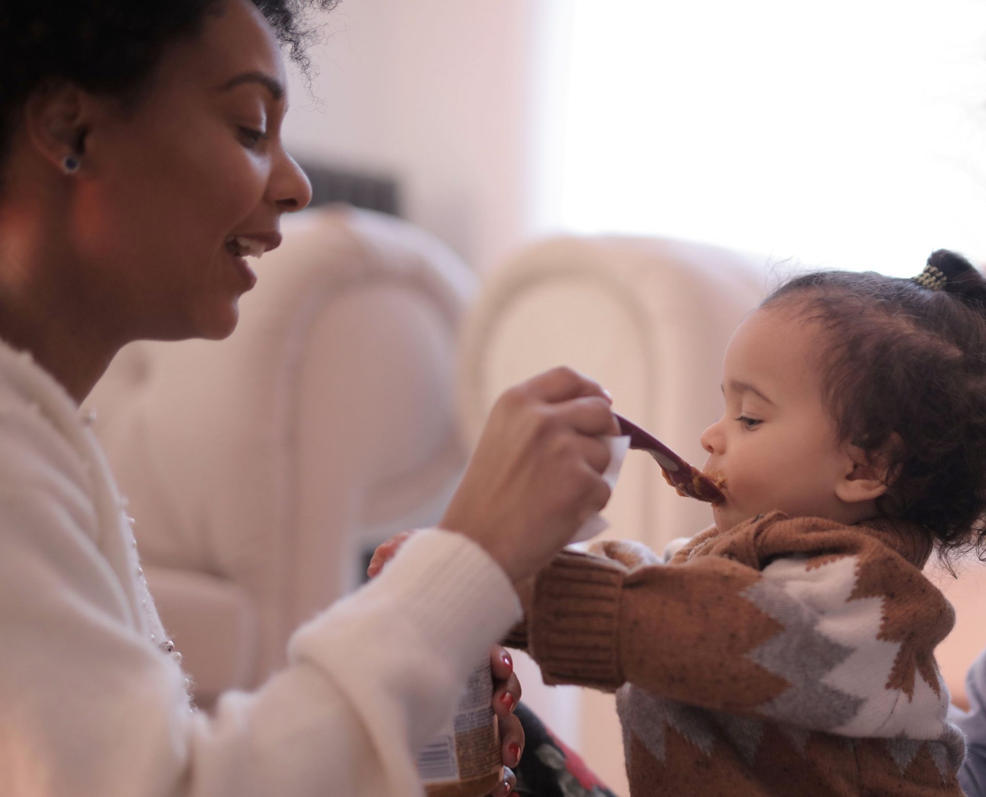 Adult feeding toddler with a spoon in a cozy living room.