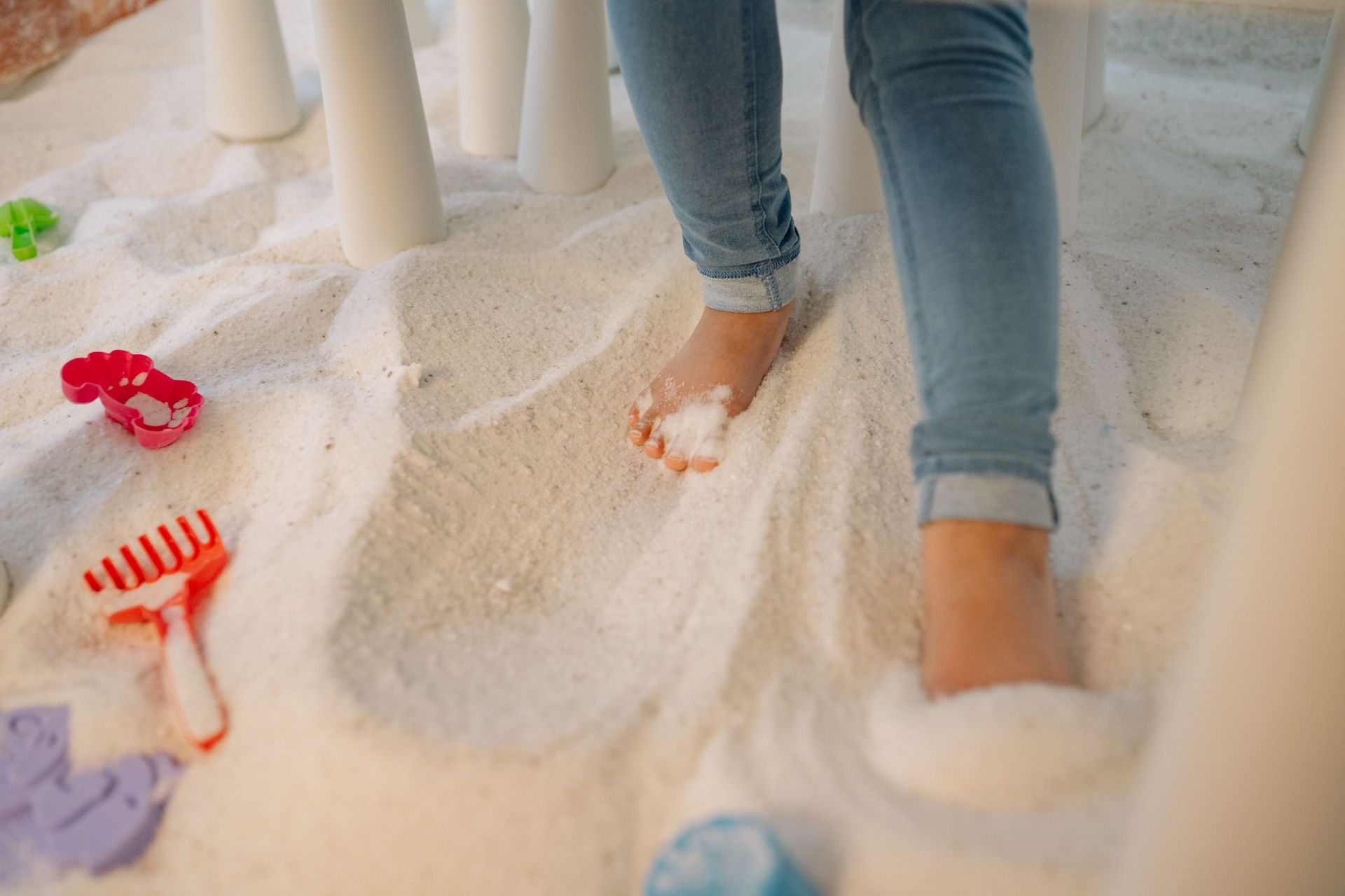 Feet in blue jeans standing in white sand with small red toys scattered nearby