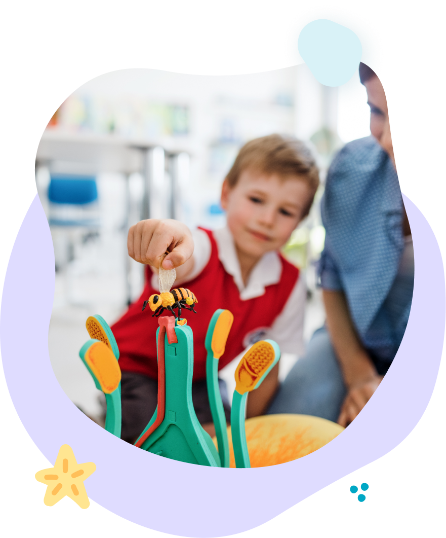 Boy playing a game with a toy bee, observed by an adult. Child is in a classroom setting.