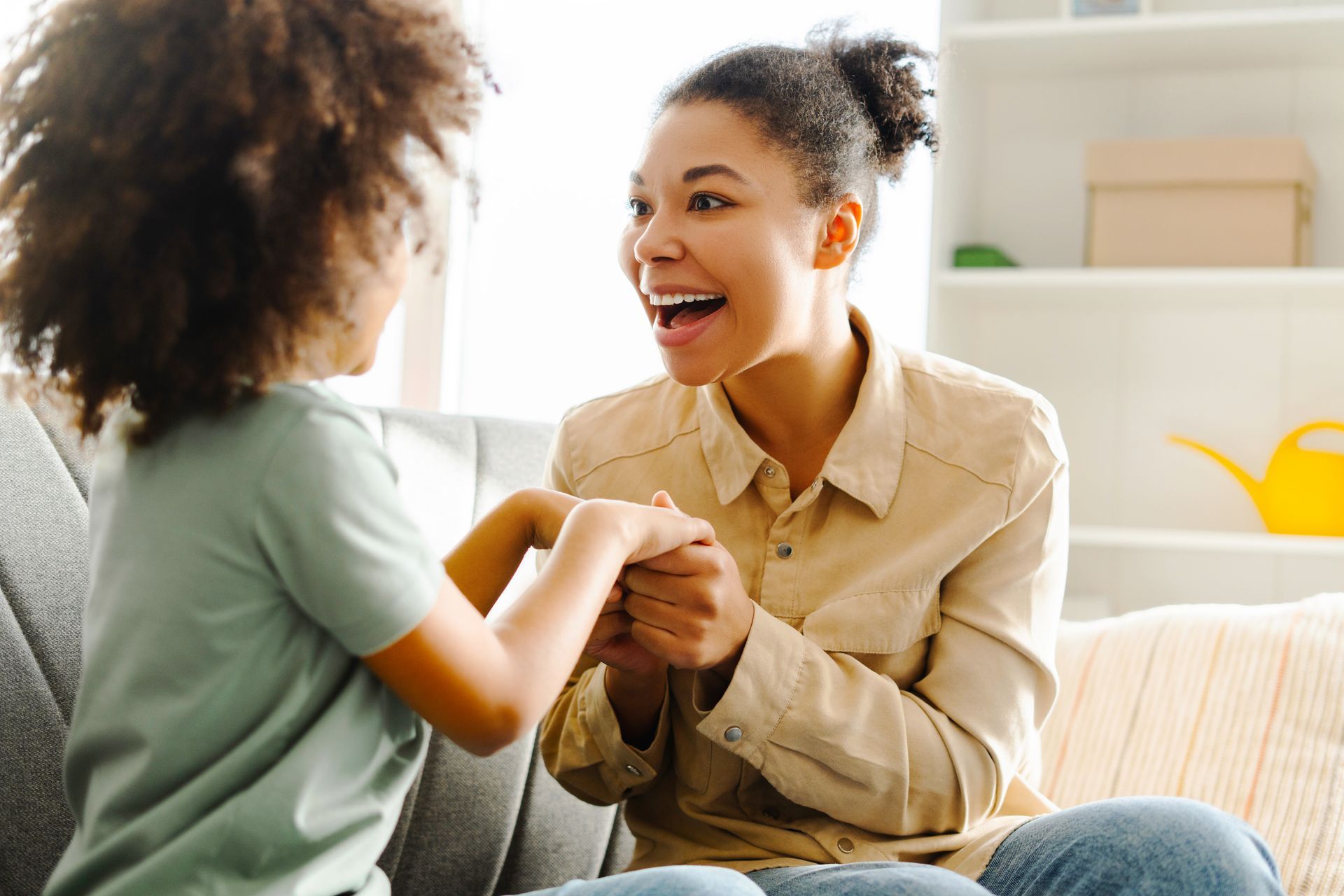 Woman and child sitting, holding hands, smiling; inside, sunny room.