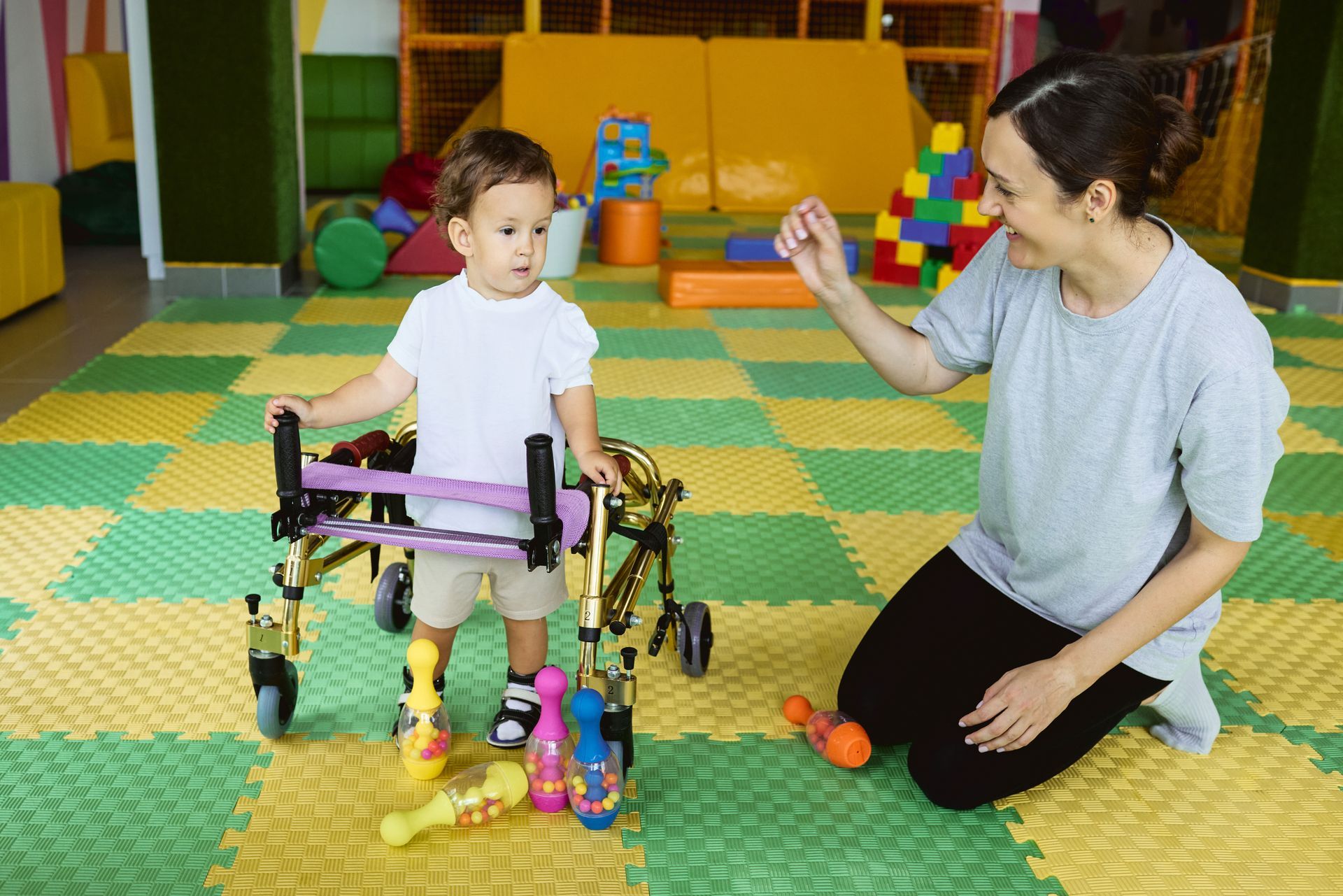 Caregiver kneels beside a child in a walker, playing with colorful toys on a padded playroom floor.