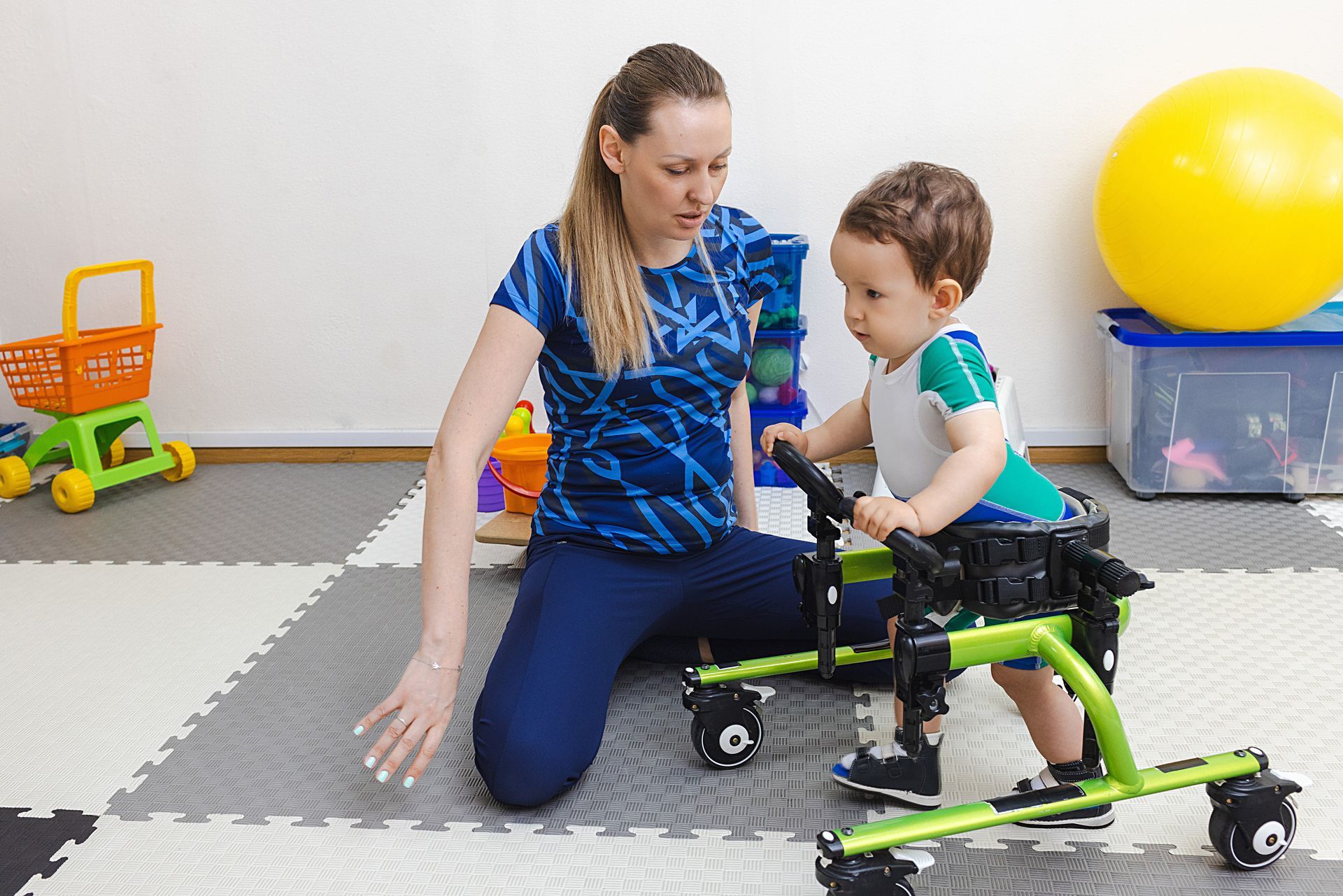 Therapist assisting a child with mobility exercises on a mat in a playroom, with toys in the background
