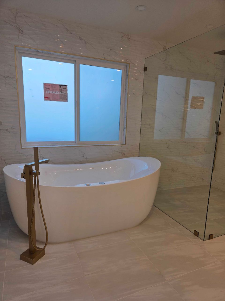 Modern bathroom with freestanding tub, glass shower, and frosted window on beige tile wall