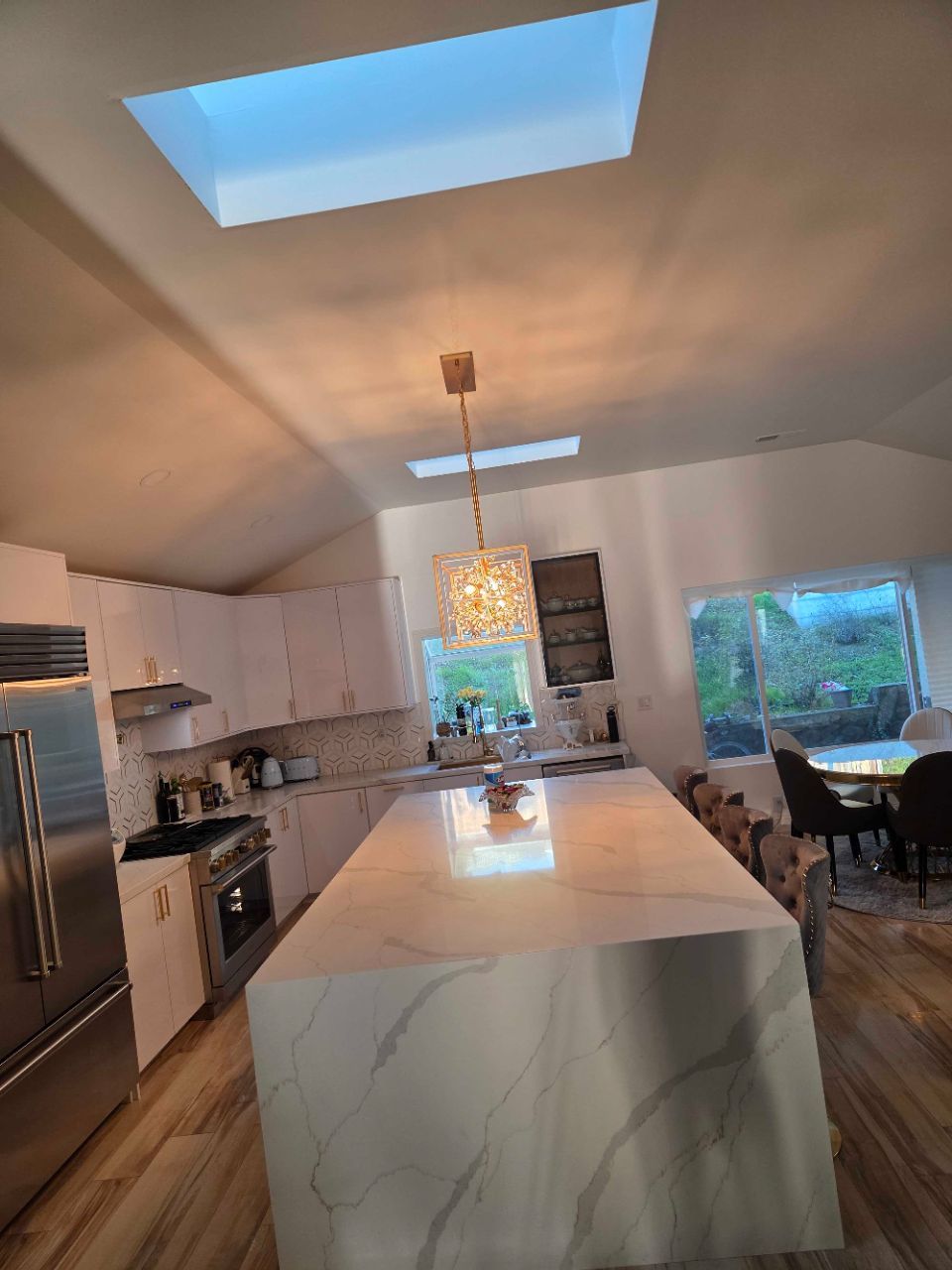 Modern kitchen with marble island, skylight, pendant light, and large windows overlooking a dining area.