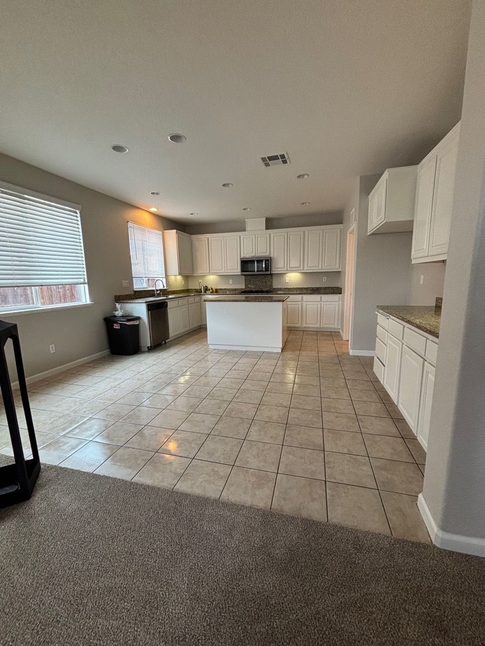 Empty open-plan kitchen and living area with tiled floor, white cabinets, and carpeted seating space