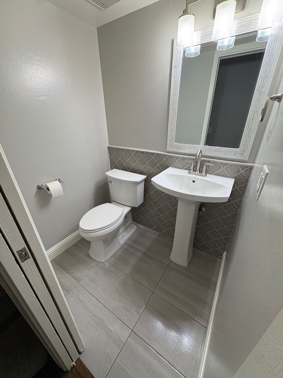 Small bathroom with toilet, pedestal sink, mirror, and gray tile floor.