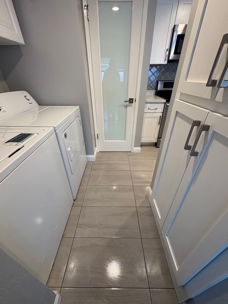 Narrow white kitchen hallway with tiled floor, cabinets on both sides, and a frosted glass door at the end