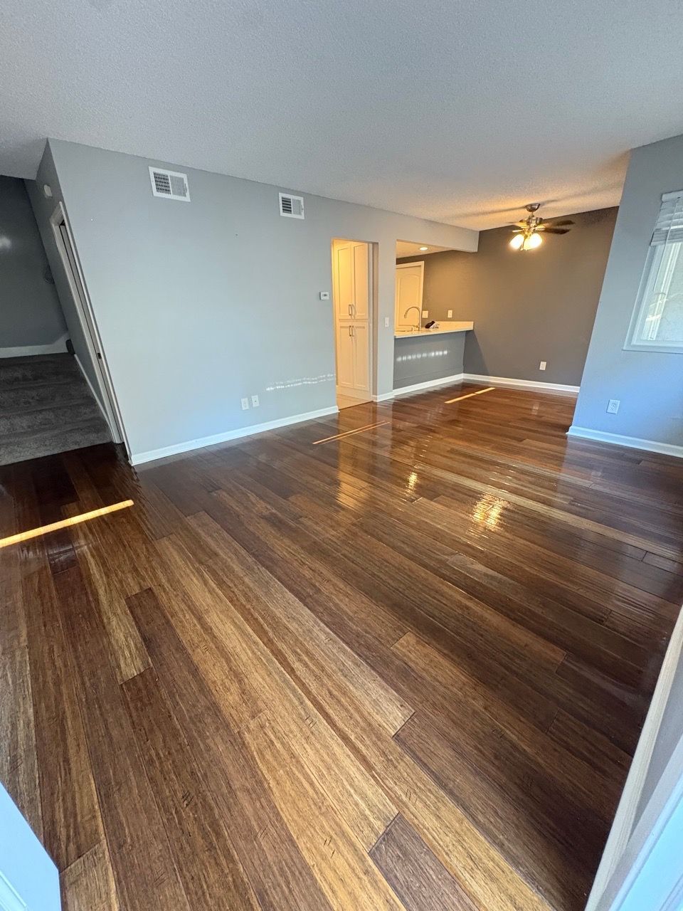Empty living room with glossy hardwood floors, gray walls, and a kitchen area in the background
