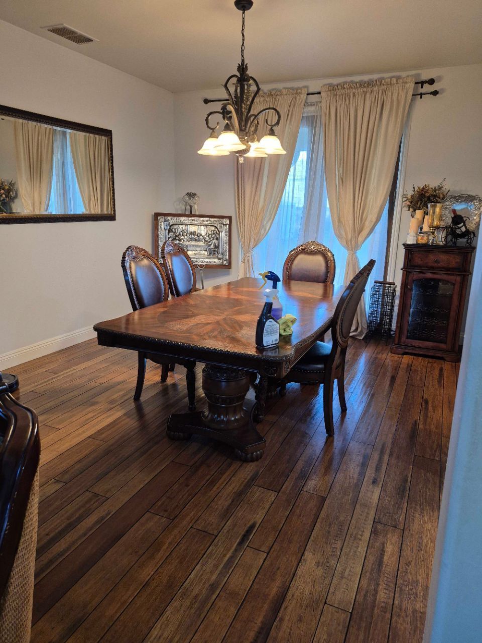 Dining room with a dark wood table, upholstered chairs, chandelier, and curtains over hardwood floors.